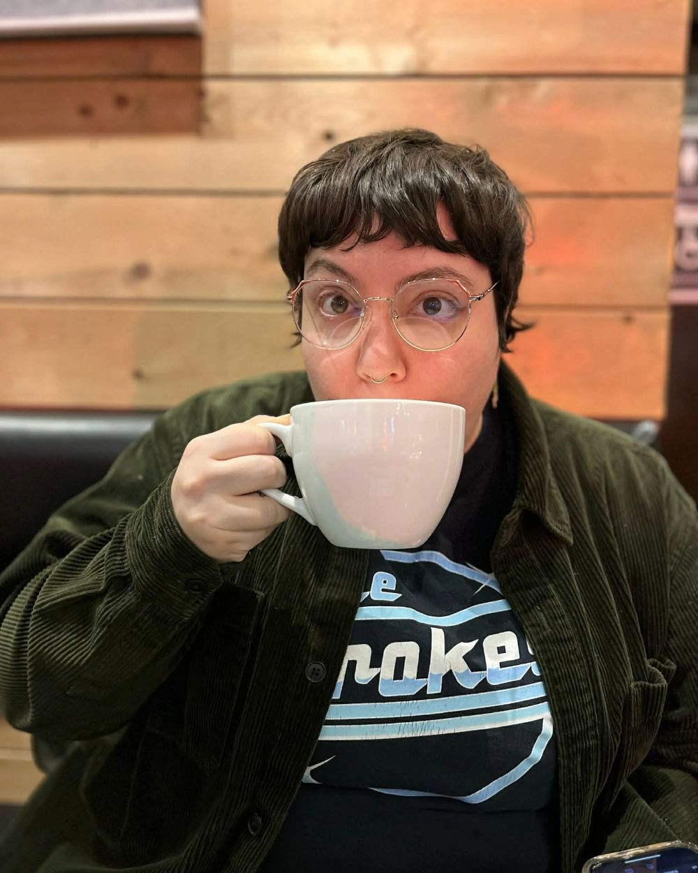 Andie sipping from a white coffee cup, wearing glasses and a corduroy shirt over a Strokes t-shirt